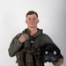 A young male military aviator is posed against a plain light gray studio background in a formal portrait-style photograph. He is wearing a full flight suit ensemble including a survival vest, harness, and gloves, and is holding a flight helmet in his right hand. The image appears to be an official or professional military portrait.