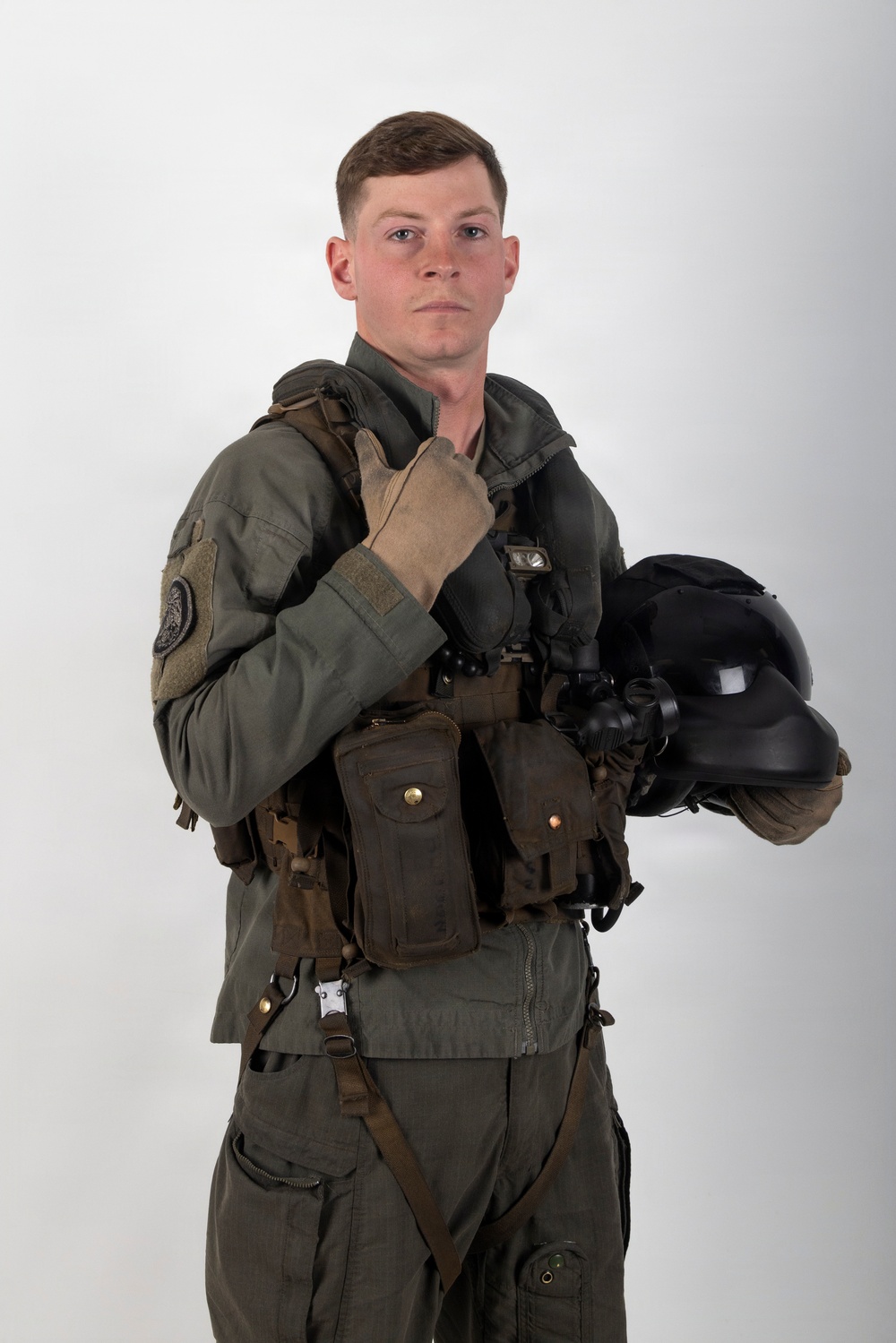 A young male military aviator poses against a plain white/light gray studio background. He is wearing a full flight suit ensemble including a survival vest, harness, and tactical gear, and is holding a flight helmet in his left arm. His right hand is raised in a thumbs-up gesture toward his chin.