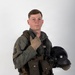 A young male military aviator poses against a plain white/light gray studio background. He is wearing a full flight suit ensemble including a survival vest, harness, and tactical gear, and is holding a flight helmet in his left arm. His right hand is raised in a thumbs-up gesture toward his chin.