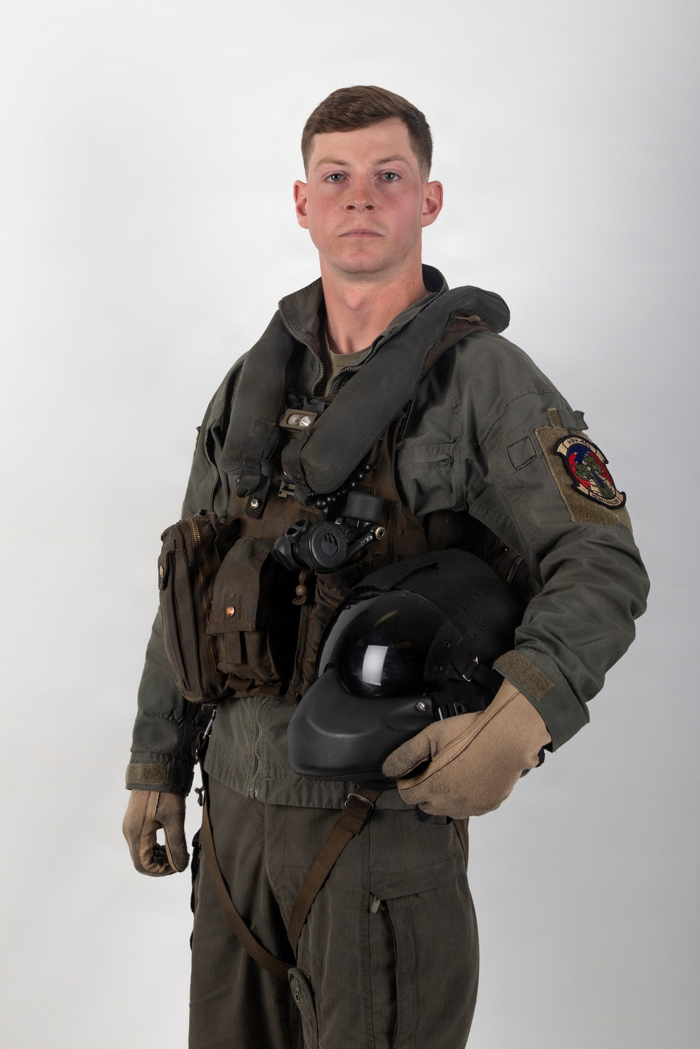 A young male military aviator poses for a formal portrait against a plain white/grey background. He is wearing a full flight suit ensemble including a green flight suit, survival vest, G-suit, and is holding a flight helmet and gloves. A squadron patch is visible on his right sleeve.