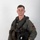 MCAS Marines pose for content 2026 A young male military aviator poses for a formal portrait against a plain white/grey background. He is wearing a full flight suit ensemble including a green flight suit, survival vest, G-suit, and is holding a flight helmet and gloves. A squadron patch is visible on his right sleeve.