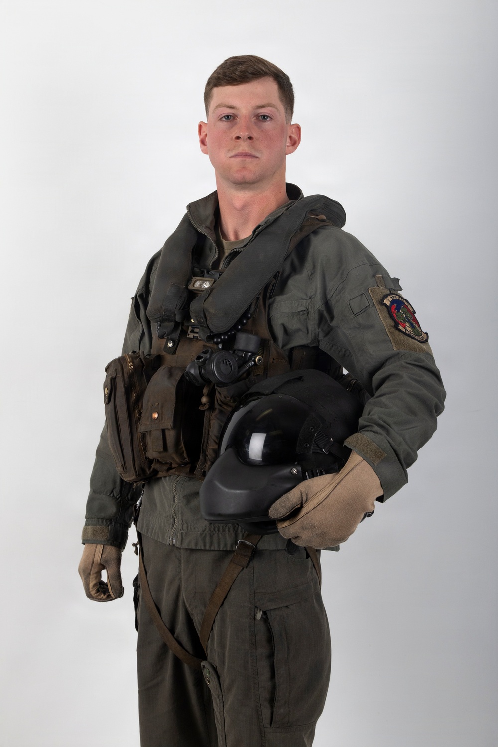 A young male military aviator is posed against a neutral white/grey studio background in a formal portrait-style photograph. He is wearing a full flight suit ensemble including a survival vest, G-suit components, and is holding a flight helmet under his right arm. A squadron patch is visible on his left sleeve.