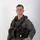 MCAS Marines pose for content 2026 A young male military aviator is posed against a neutral white/grey studio background in a formal portrait-style photograph. He is wearing a full flight suit ensemble including a survival vest, G-suit components, and is holding a flight helmet under his right arm. A squadron patch is visible on his left sleeve.
