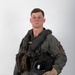 A young male military aviator is posed against a neutral white/grey studio background in a formal portrait-style photograph. He is wearing a full flight suit ensemble including a survival vest, G-suit components, and is holding a flight helmet under his right arm. A squadron patch is visible on his left sleeve.