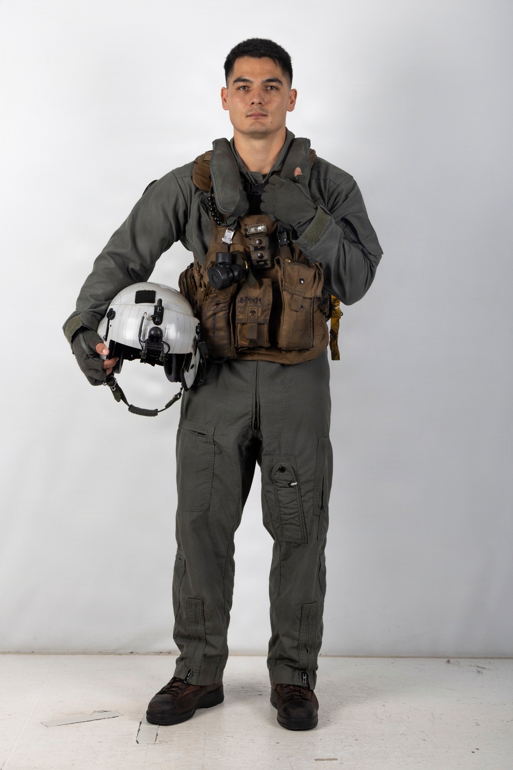 A young male military aviator stands in a full-body portrait pose against a plain white/light gray studio background. He is wearing a complete flight suit ensemble including a sage/olive green flight suit, tactical chest rig, and is holding a white flight helmet. His right hand is raised in a thumbs-up gesture.