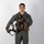 MCAS Marines pose for content 2026 A young male military aviator stands in a full-body portrait pose against a plain white/light gray studio background. He is wearing a complete flight suit ensemble including a sage/olive green flight suit, tactical chest rig, and is holding a white flight helmet. His right hand is raised in a thumbs-up gesture.