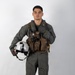 A young male military aviator stands in a full-body portrait pose against a plain white/light gray studio background. He is wearing a complete flight suit ensemble including a sage/olive green flight suit, tactical chest rig, and is holding a white flight helmet. His right hand is raised in a thumbs-up gesture.
