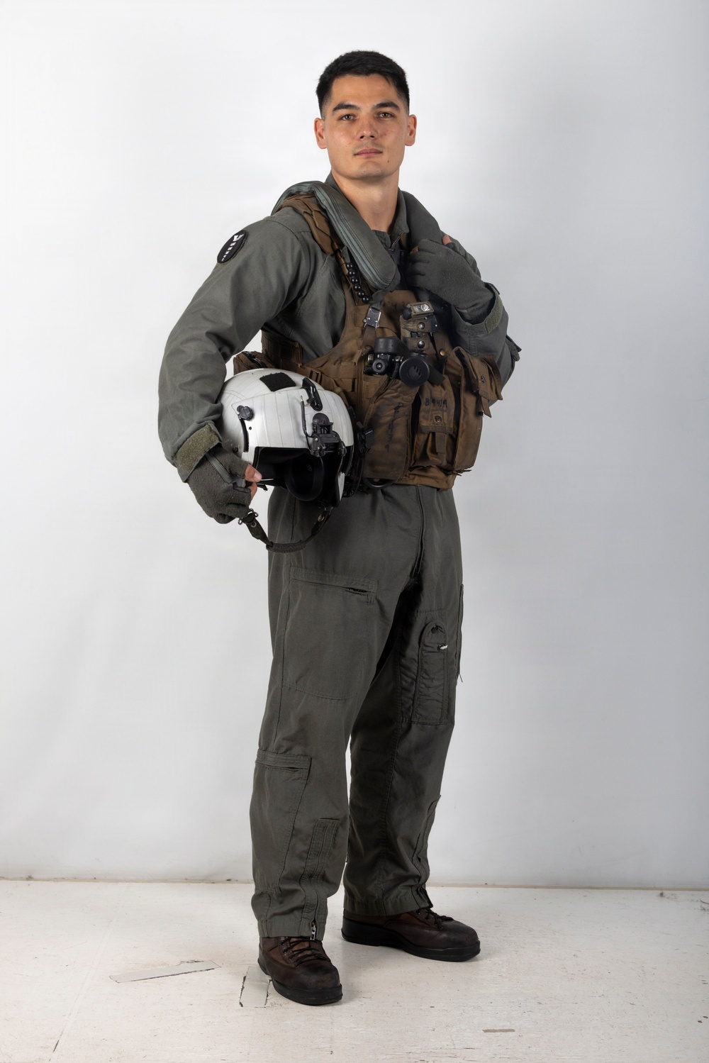 A young male military aviator stands in a full-body portrait pose against a plain white background. He is wearing a olive-green flight suit with a tactical survival vest and is holding a white flight helmet under his left arm. The image appears to be a formal or official military portrait photograph.