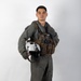 A young male military aviator stands in a full-body portrait pose against a plain white background. He is wearing a olive-green flight suit with a tactical survival vest and is holding a white flight helmet under his left arm. The image appears to be a formal or official military portrait photograph.