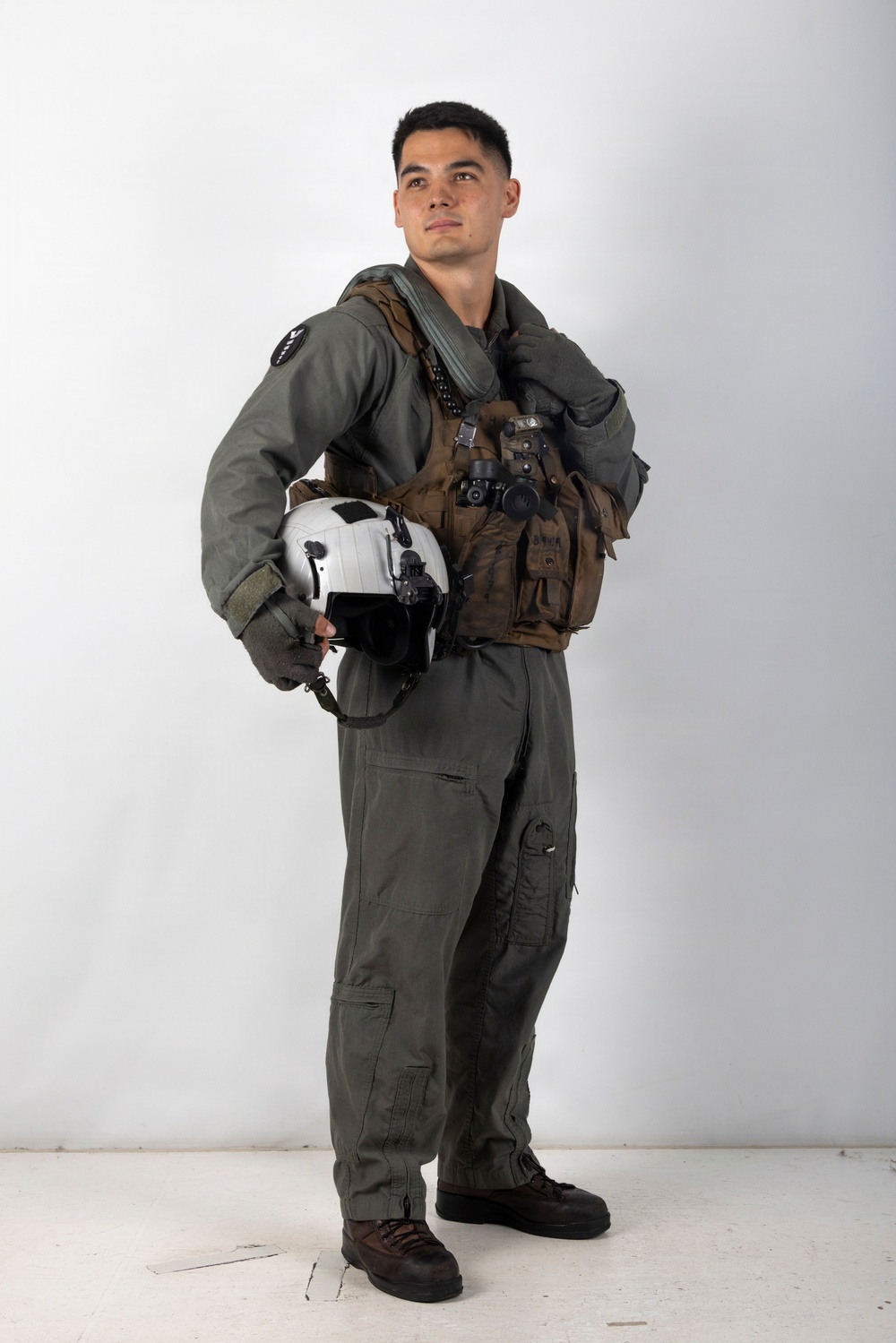 A young male military aviator stands in a full-body portrait pose against a plain white background. He is wearing a sage/olive green flight suit with a tactical survival vest and holds a white flight helmet at his side. The image appears to be a professional or semi-professional portrait photograph.