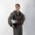 MCAS Marines pose for content 2026 A young male military aviator stands in a full-body portrait pose against a plain white background. He is wearing a sage/olive green flight suit with a tactical survival vest and holds a white flight helmet at his side. The image appears to be a professional or semi-professional portrait photograph.