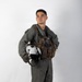 A young male military aviator stands in a full-body portrait pose against a plain white background. He is wearing a sage/olive green flight suit with a tactical survival vest and holds a white flight helmet at his side. The image appears to be a professional or semi-professional portrait photograph.
