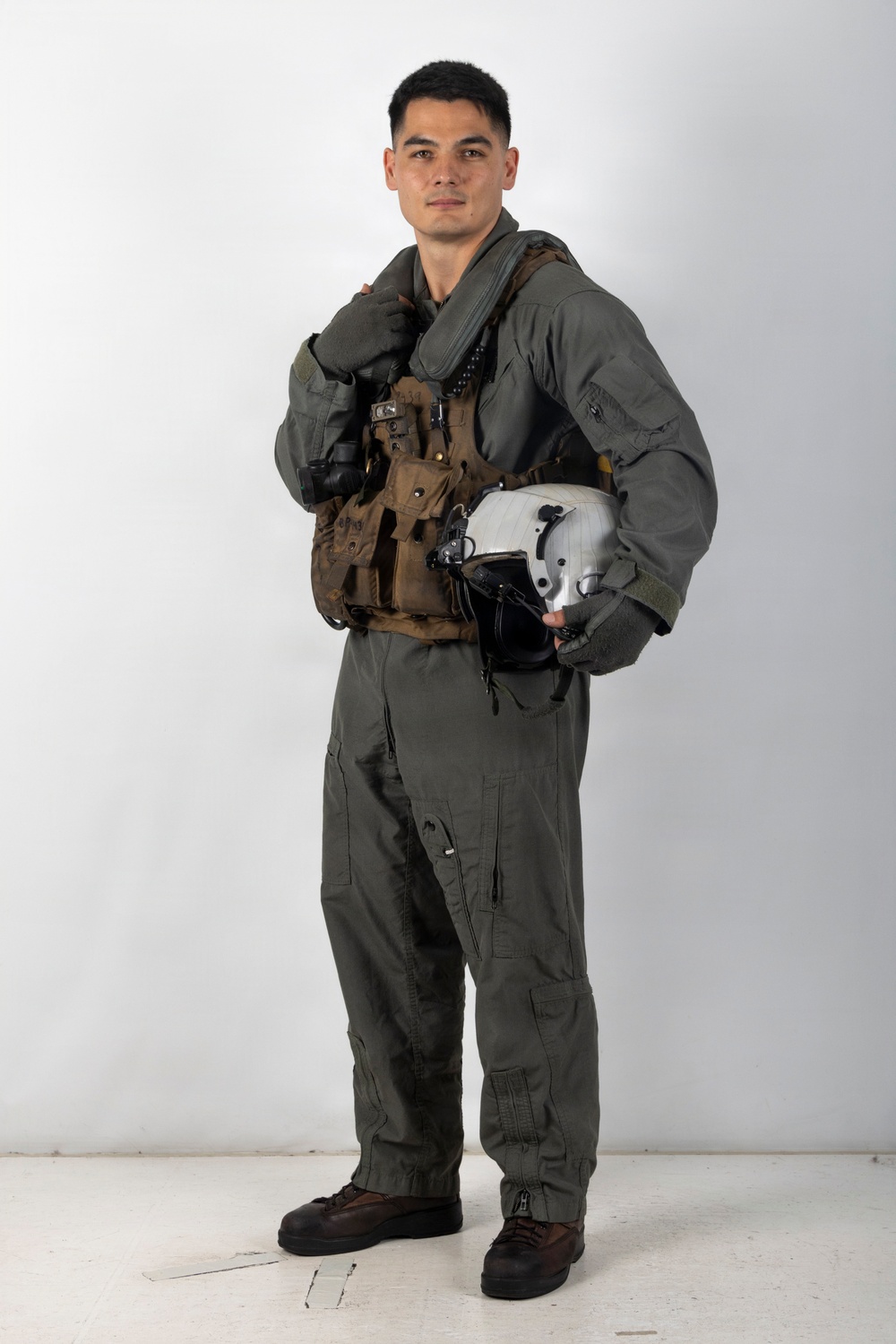 A young male military aviator is posed against a plain white/light gray studio backdrop in a full-length portrait. He is wearing an olive-drab flight suit with a tactical survival vest and is holding a white flight helmet under his right arm. The image appears to be an official or promotional military portrait.