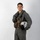 MCAS Marines pose for content 2026 A young male military aviator is posed against a plain white/light gray studio backdrop in a full-length portrait. He is wearing an olive-drab flight suit with a tactical survival vest and is holding a white flight helmet under his right arm. The image appears to be an official or promotional military portrait.