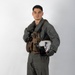 A young male military aviator is posed against a plain white/light gray studio backdrop in a full-length portrait. He is wearing an olive-drab flight suit with a tactical survival vest and is holding a white flight helmet under his right arm. The image appears to be an official or promotional military portrait.