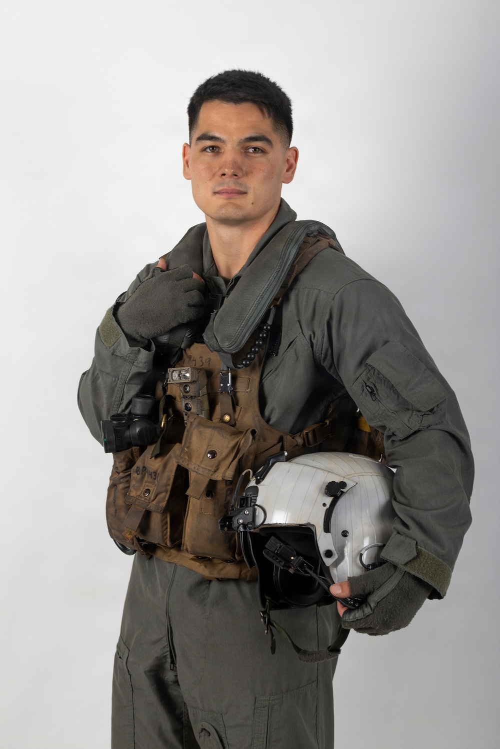 A young male military aviator poses against a plain white/light gray studio background. He is wearing a full flight suit ensemble including a sage green flight suit, tactical survival vest, and fingerless flight gloves, and is holding a white flight helmet. The image appears to be an official or semi-official portrait-style photograph.