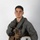 MCAS Marines pose for content 2026 A young male military aviator poses against a plain white/light gray studio background. He is wearing a full flight suit ensemble including a sage green flight suit, tactical survival vest, and fingerless flight gloves, and is holding a white flight helmet. The image appears to be an official or semi-official portrait-style photograph.