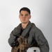 A young male military aviator poses against a plain white/light gray studio background. He is wearing a full flight suit ensemble including a sage green flight suit, tactical survival vest, and fingerless flight gloves, and is holding a white flight helmet. The image appears to be an official or semi-official portrait-style photograph.