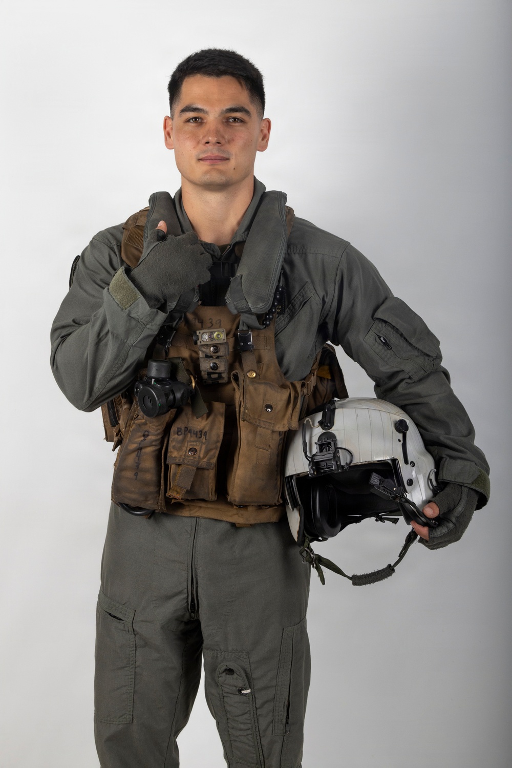 A young male military aviator poses against a neutral gray studio background in full flight gear. He is holding a flight helmet under his right arm and giving a thumbs-up gesture with his left hand. The equipment appears well-used and bears handwritten markings.