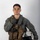 MCAS Marines pose for content 2026 A young male military aviator poses against a neutral gray studio background in full flight gear. He is holding a flight helmet under his right arm and giving a thumbs-up gesture with his left hand. The equipment appears well-used and bears handwritten markings.