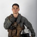 A young male military aviator poses against a neutral gray studio background in full flight gear. He is holding a flight helmet under his right arm and giving a thumbs-up gesture with his left hand. The equipment appears well-used and bears handwritten markings.