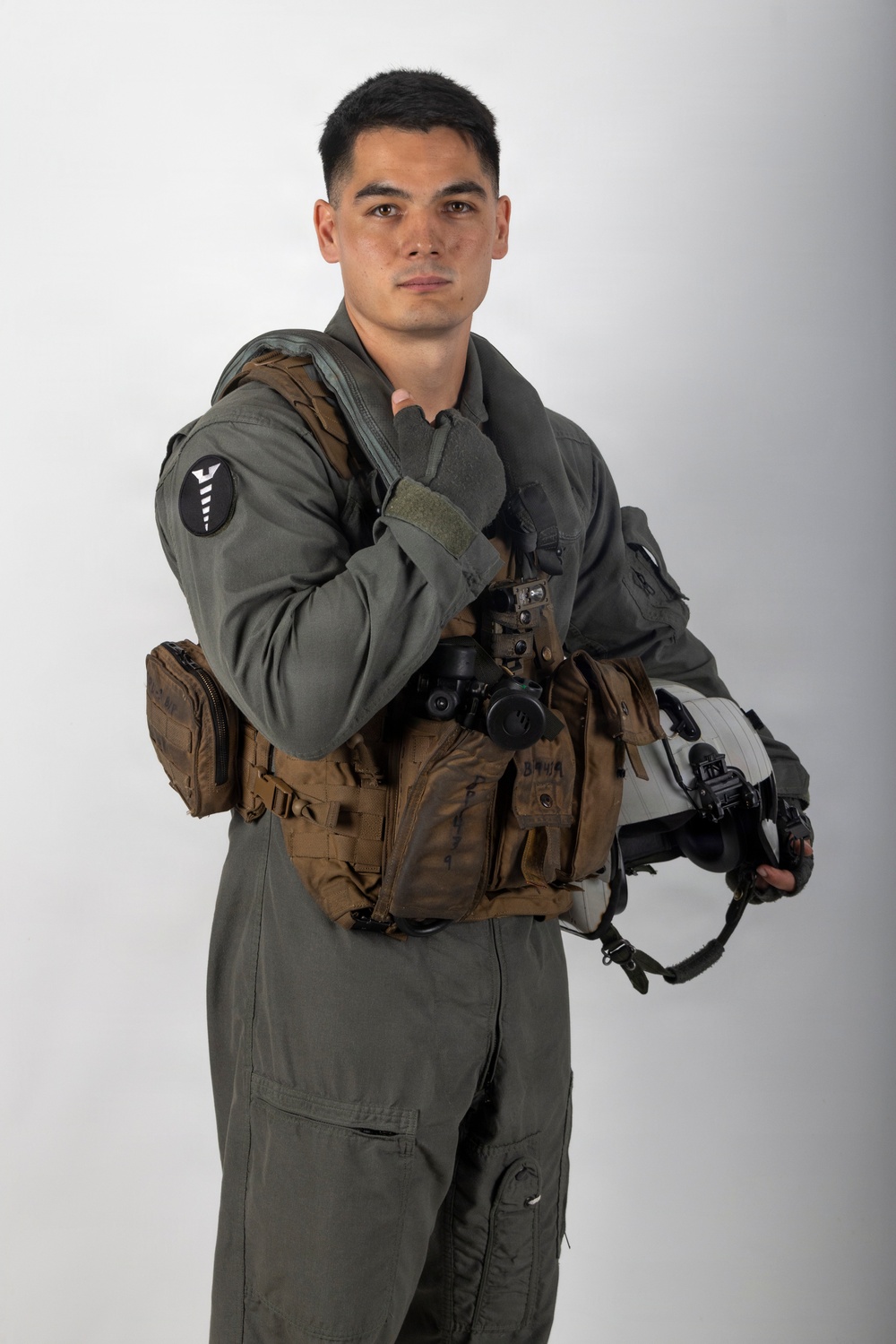 A young male military aviator poses against a neutral gray-white studio background in a formal portrait-style photograph. He is wearing a sage/olive green flight suit with a tactical survival vest and is holding a flight helmet. The image appears to be an official or semi-official military portrait.