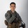 MCAS Marines pose for content 2026 A young male military aviator poses against a neutral gray-white studio background in a formal portrait-style photograph. He is wearing a sage/olive green flight suit with a tactical survival vest and is holding a flight helmet. The image appears to be an official or semi-official military portrait.