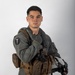 A young male military aviator poses against a neutral gray-white studio background in a formal portrait-style photograph. He is wearing a sage/olive green flight suit with a tactical survival vest and is holding a flight helmet. The image appears to be an official or semi-official military portrait.