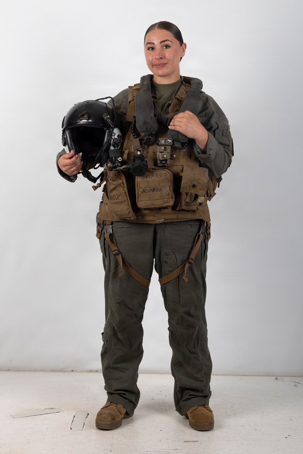 A female military aviator stands in a full-body portrait against a white studio background. She is wearing a complete flight suit ensemble including a survival vest, harness, and is holding a flight helmet. The image appears to be an official or professional military portrait.