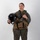 MCAS Marines pose for content 2026 A female military aviator stands in a full-body portrait against a white studio background. She is wearing a complete flight suit ensemble including a survival vest, harness, and is holding a flight helmet. The image appears to be an official or professional military portrait.