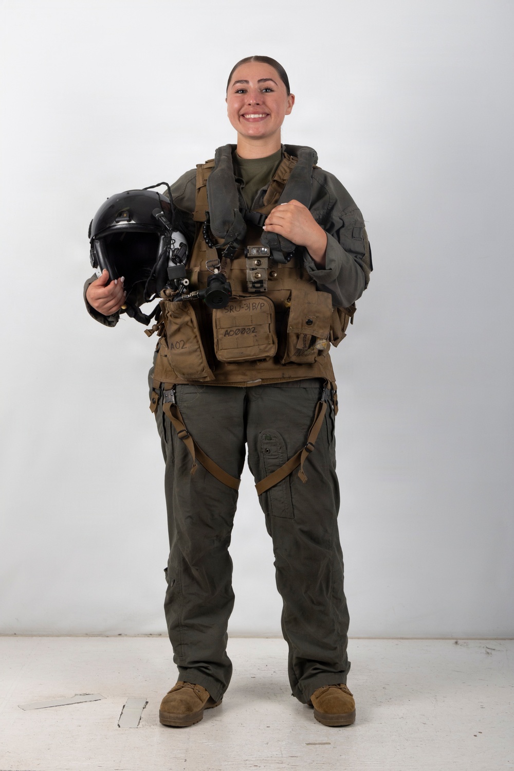 A smiling individual stands in full military flight gear against a plain white background in what appears to be an official portrait-style photograph. They are wearing an olive/sage green flight suit with a tactical survival vest labeled 'SRU-31B/P' and holding a black flight helmet with visor and oxygen mask attachment. The overall composition and equipment suggest a U.S. military aviator in standard flight crew gear.