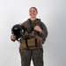 A smiling individual stands in full military flight gear against a plain white background in what appears to be an official portrait-style photograph. They are wearing an olive/sage green flight suit with a tactical survival vest labeled 'SRU-31B/P' and holding a black flight helmet with visor and oxygen mask attachment. The overall composition and equipment suggest a U.S. military aviator in standard flight crew gear.