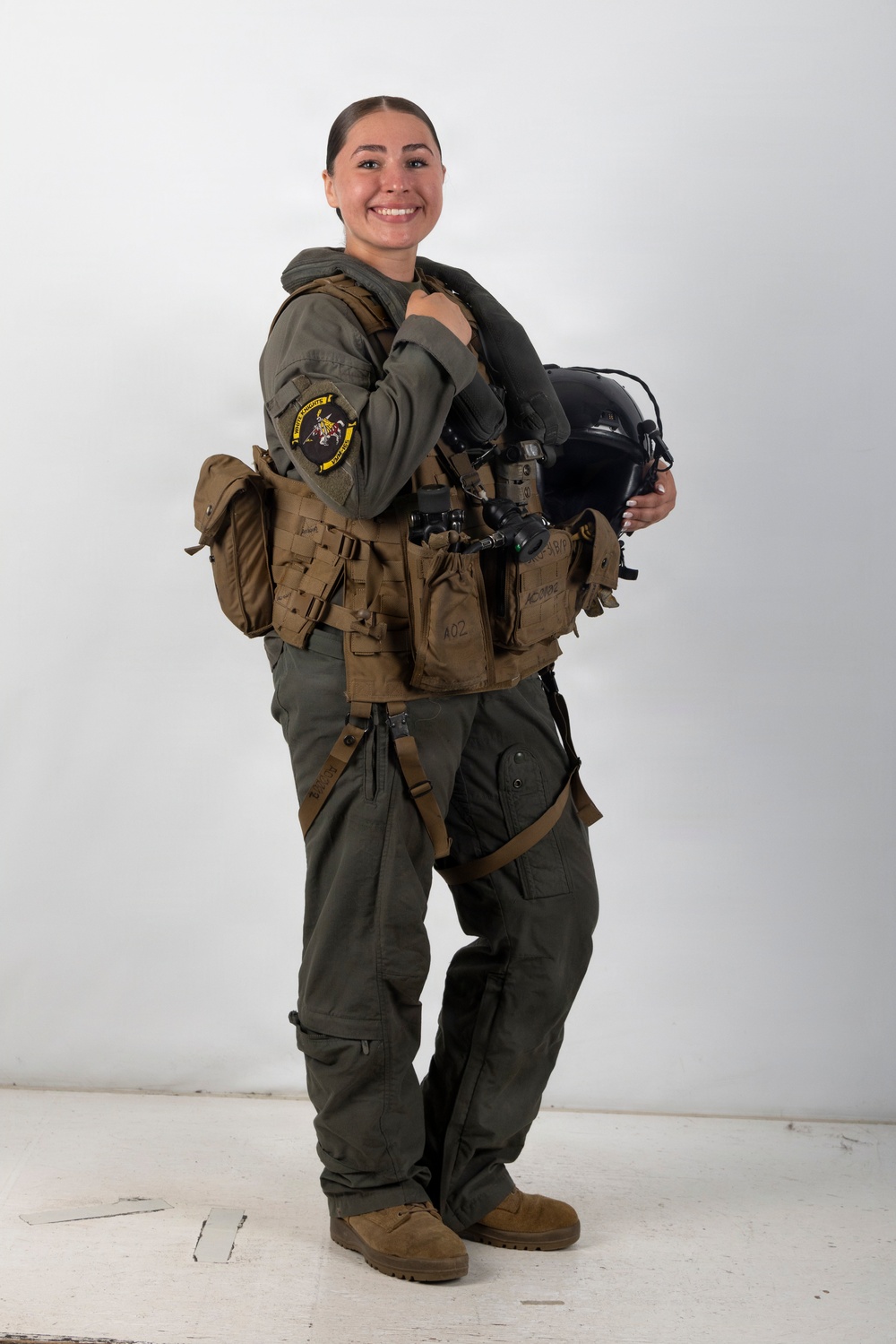 A smiling female military aviator poses in full flight gear against a white studio background. She is wearing an olive green flight suit with a squadron patch on the left sleeve, a tactical survival vest, and is holding a flight helmet. The image appears to be an official or professional portrait photograph.