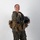 MCAS Marines pose for content 2026 A smiling female military aviator poses in full flight gear against a white studio background. She is wearing an olive green flight suit with a squadron patch on the left sleeve, a tactical survival vest, and is holding a flight helmet. The image appears to be an official or professional portrait photograph.