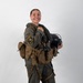 A smiling female military aviator poses in full flight gear against a white studio background. She is wearing an olive green flight suit with a squadron patch on the left sleeve, a tactical survival vest, and is holding a flight helmet. The image appears to be an official or professional portrait photograph.