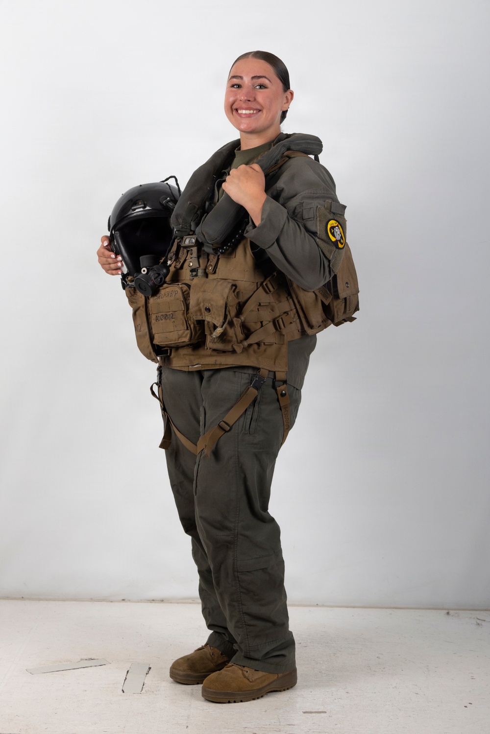 A smiling military aviator stands in a studio-style setting wearing full flight gear including a survival vest and flight suit. She is holding a black flight helmet with oxygen mask attachment in her left hand while her right hand rests on her shoulder. A yellow and black squadron patch is visible on her right sleeve.