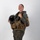 MCAS Marines pose for content 2026 A smiling military aviator stands in a studio-style setting wearing full flight gear including a survival vest and flight suit. She is holding a black flight helmet with oxygen mask attachment in her left hand while her right hand rests on her shoulder. A yellow and black squadron patch is visible on her right sleeve.