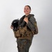 A smiling military aviator stands in a studio-style setting wearing full flight gear including a survival vest and flight suit. She is holding a black flight helmet with oxygen mask attachment in her left hand while her right hand rests on her shoulder. A yellow and black squadron patch is visible on her right sleeve.