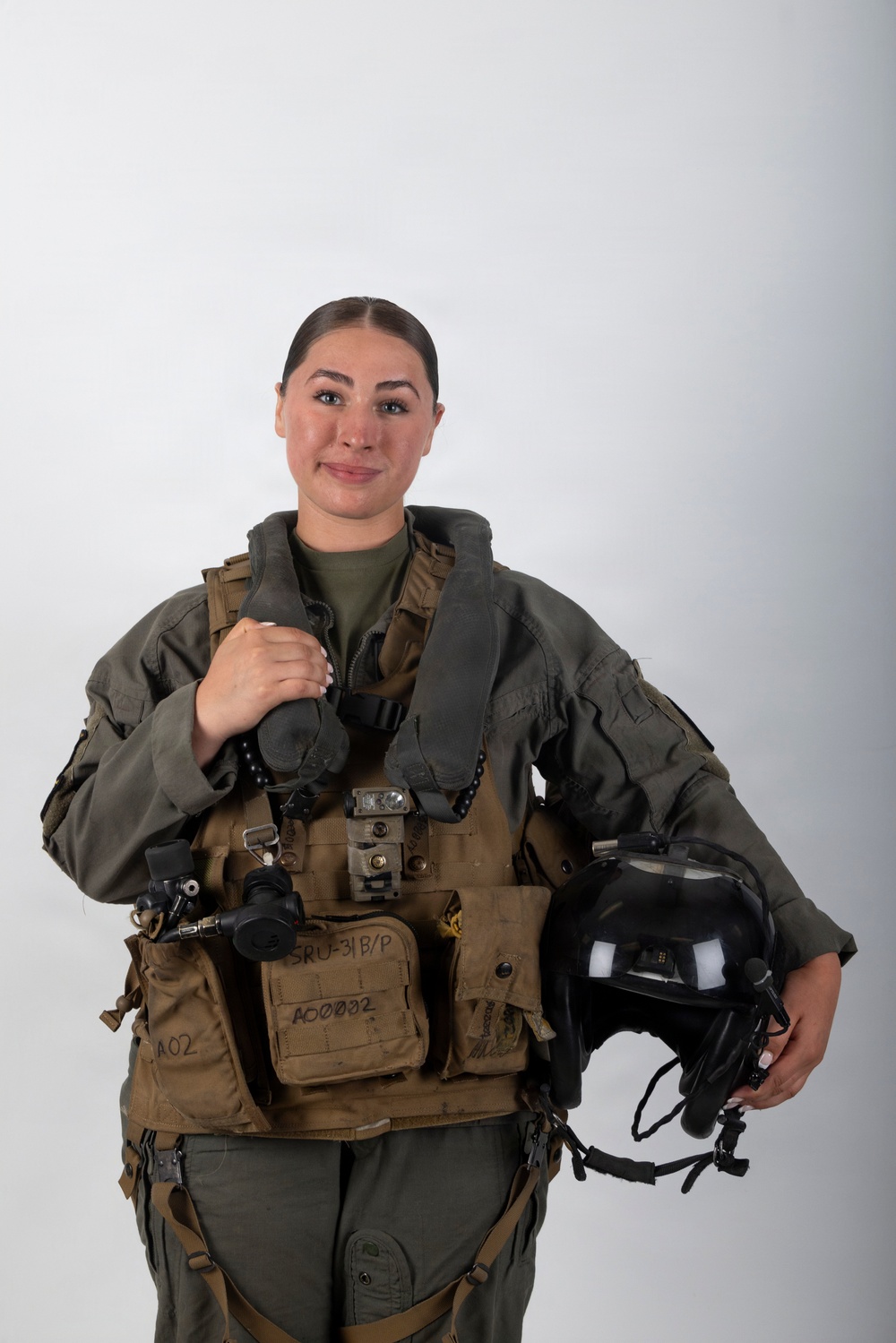 A young woman is posed in a professional portrait setting wearing full military flight gear, including a survival vest and flight suit. She is holding a flight helmet in her right hand and smiling at the camera against a plain light gray background. The equipment markings suggest U.S. military aviation affiliation.