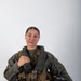 A young woman is posed in a professional portrait setting wearing full military flight gear, including a survival vest and flight suit. She is holding a flight helmet in her right hand and smiling at the camera against a plain light gray background. The equipment markings suggest U.S. military aviation affiliation.