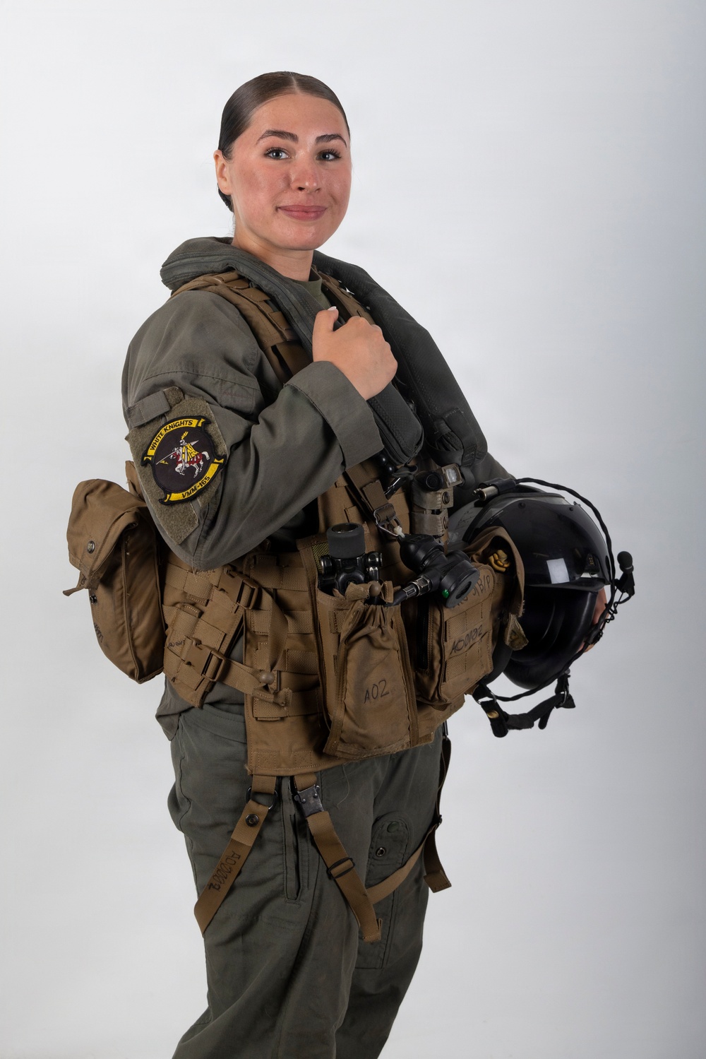 A female military aviator poses in a studio setting wearing full flight gear including a tactical vest, survival equipment, and holding a flight helmet. She is dressed in an olive green flight suit with a squadron patch visible on her left sleeve reading 'White Knights VMM-465'. The image appears to be an official or professional portrait photograph.