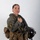 MCAS Marines pose for content 2026 A female military aviator poses in a studio setting wearing full flight gear including a tactical vest, survival equipment, and holding a flight helmet. She is dressed in an olive green flight suit with a squadron patch visible on her left sleeve reading 'White Knights VMM-465'. The image appears to be an official or professional portrait photograph.