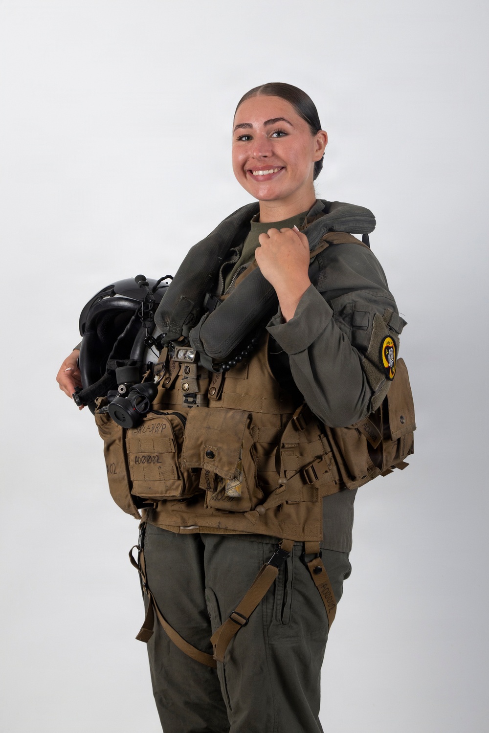 A smiling individual is posed against a plain white background wearing full military aviation flight gear, including a tactical vest and flight suit. They are holding a flight helmet with an attached oxygen mask and night vision goggle mount. The image appears to be a professional portrait or official military photo.