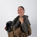 A smiling individual is posed against a plain white background wearing full military aviation flight gear, including a tactical vest and flight suit. They are holding a flight helmet with an attached oxygen mask and night vision goggle mount. The image appears to be a professional portrait or official military photo.