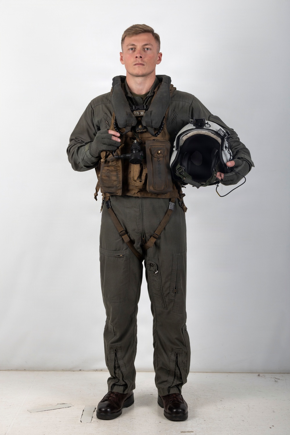A young male military aviator stands in a formal portrait pose against a plain white/grey studio background. He is wearing a full flight suit ensemble with survival vest and harness, holding a flight helmet in his right hand. The image appears to be an official or professional military portrait photograph.