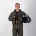 A young male military aviator stands in a formal portrait pose against a plain white/grey studio background. He is wearing a full flight suit ensemble with survival vest and harness, holding a flight helmet in his right hand. The image appears to be an official or professional military portrait photograph.