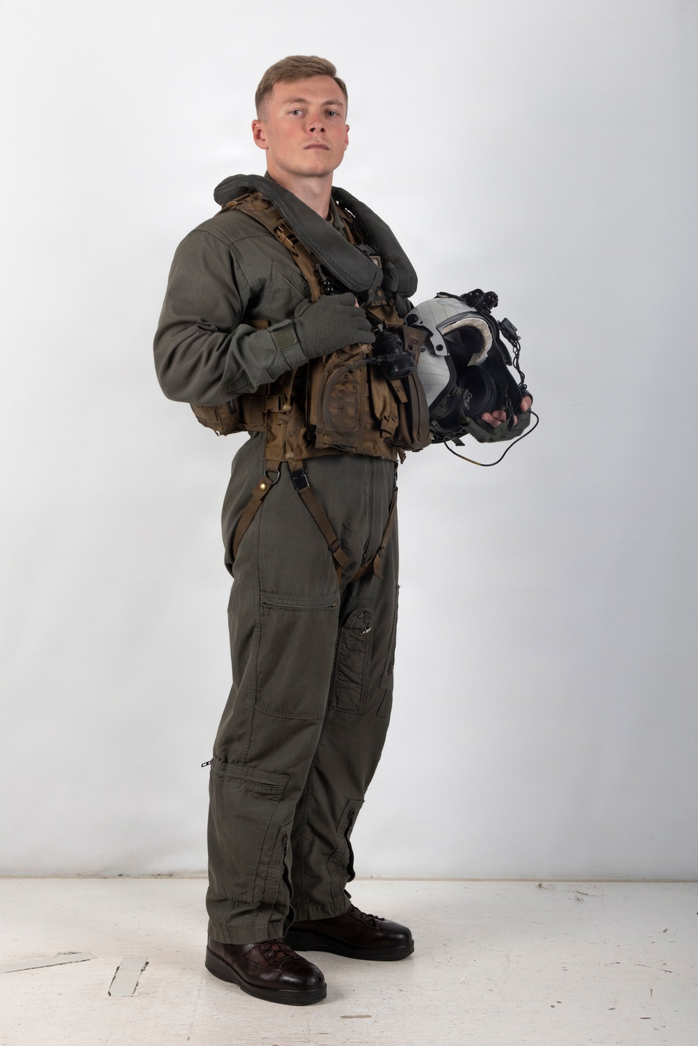 A young male military aviator stands in a full-body portrait pose against a plain white/light gray studio backdrop. He is wearing an olive drab flight suit with full tactical gear including a survival vest, harness, and inflatable collar. He holds a flight helmet with attached oxygen mask and night vision mount in his left hand.