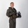 MCAS Marines pose for content 2026 A young male military aviator stands in a full-body portrait pose against a plain white/light gray studio backdrop. He is wearing an olive drab flight suit with full tactical gear including a survival vest, harness, and inflatable collar. He holds a flight helmet with attached oxygen mask and night vision mount in his left hand.