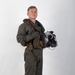 A young male military aviator stands in a full-body portrait pose against a plain white/light gray studio backdrop. He is wearing an olive drab flight suit with full tactical gear including a survival vest, harness, and inflatable collar. He holds a flight helmet with attached oxygen mask and night vision mount in his left hand.