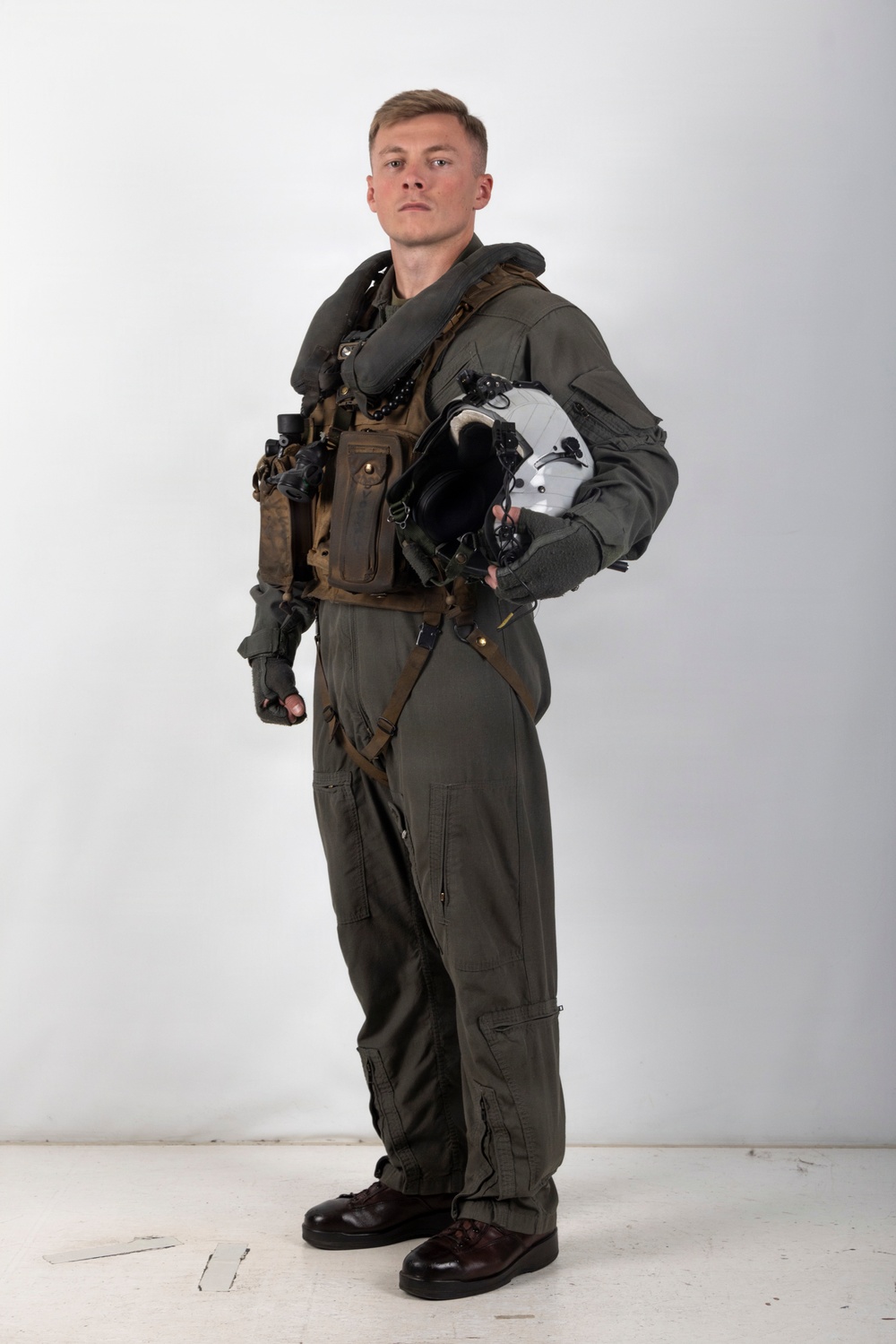 A young male military aviator stands in a full-body portrait pose against a plain white/light grey studio background. He is wearing a complete flight suit ensemble including a green/olive flight suit, survival vest, harness, and neck collar, and is holding a white flight helmet under his arm. The image appears to be an official or professional military portrait photograph.
